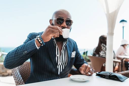 A portrait of a dapper bald African man in sunglasses and a fashionable summer suit sitting in a street cafe and drinking coffee, with a selective focus on his hand with a cup of espresso
