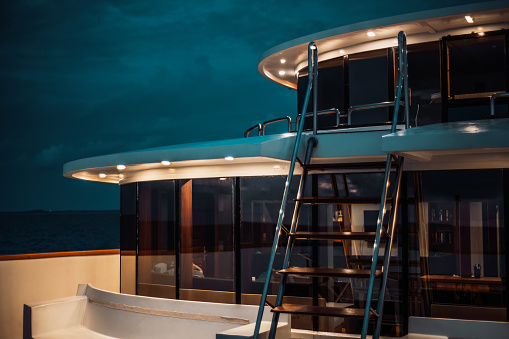 A night low-key shot of a luxury diving safari yacht deck lit by multiple ceiling lamps, with a ladder in front and a glass row of windows with a wardroom behind it; a twilight sky in the background