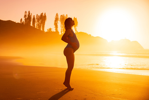 Pregnant woman in swimwear posing on beach with bright sunrise or sunset tones and trees on background