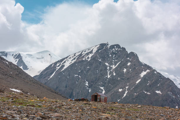 pequeña cabaña a gran altitud cerca del borde del precipicio con vista a la alta montaña rocosa con cima de pico. pequeña casa contra pico puntiagudo nevado bajo cielo nublado. rusia, república de altái, aktru, 06 de julio de 2021. - rocky mountian fotografías e imágenes de stock