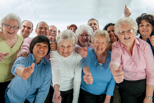 A large group of diverse seniors pose together for a portrait.  They are each dressed casually and have their thumbs up as they smile for the camera.