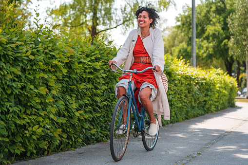 Multiracial woman with curly, black hair, riding a bike on a pavement,  looking happy, her skirt pulled up over the knees. Full length shot, bright daylight and contrasting shadow. Green in the background.
