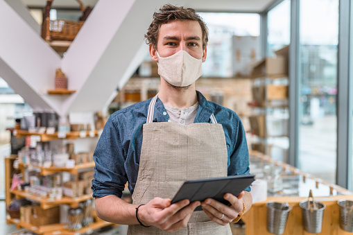 Portrait of a young adult male zero waste shop employee wearing protective face mask using a digital tablet. Plastic free sustainable environment. He is wearing an apron.