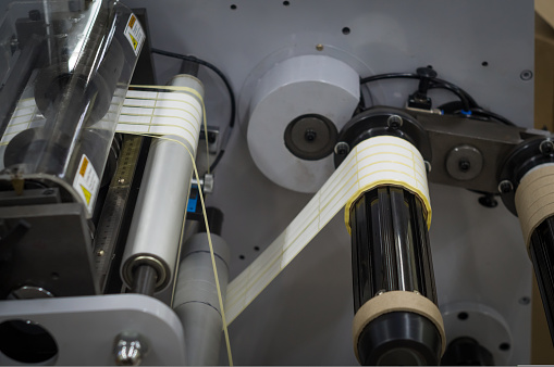 A machine for the production of white self-adhesive labels. The shafts of the machine for winding rolls of tape with a label. Die-cutting and cutting of paper for label production. Selective focus.