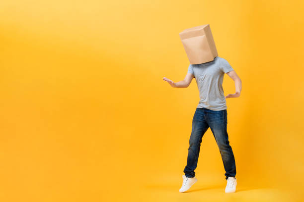 Anonymous man with head covered with paper bag opening hands in yellow studio isolated background with copy space stock photo