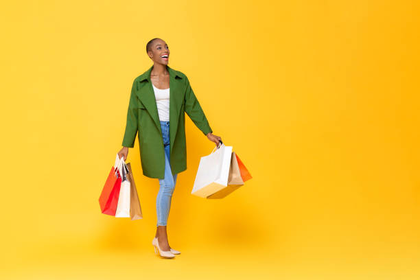 黄色のスタジオの孤立した背景の上を歩くカラフルなショッピングバッグを運ぶトレンディなファッショナブルなアフリカ系アメリカ人女性 - carry clothes ストックフォトと画像