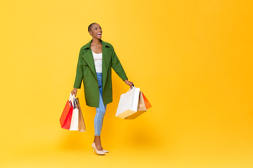 Trendy fashionable African American woman carrying colorful shopping bags walking on yellow color studio isolated background