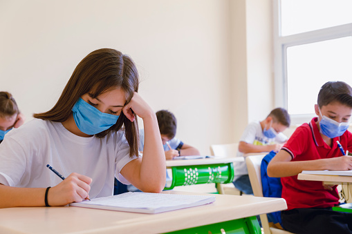 Little high school student boy wearing protective medical facemask sitting, studying in classroom completing assignment in class with their classmates taking notes from book due to coronavirus emergency Covid-19 pandemic.