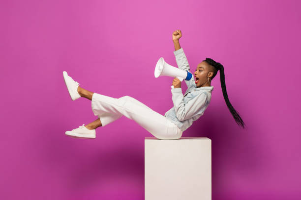 молодая афроамериканка кричит на мегафон и поднимает кулак в студии фиолетово-розового цвета на изолированном фоне - announcement message megaphone public speaker women стоковые фото и изображения