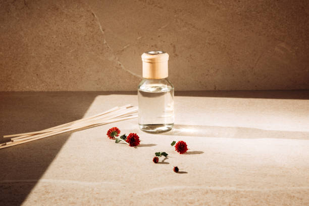 Aroma diffuser in a clear glass bottle with a wooden cap on a light stone background with wooden sticks behind and red chrysanthemum flowers