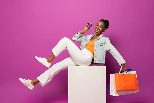 Trendy fashionable African American woman holding colorful shopping bags and credit card sitting on stool in pink color studio isolated background