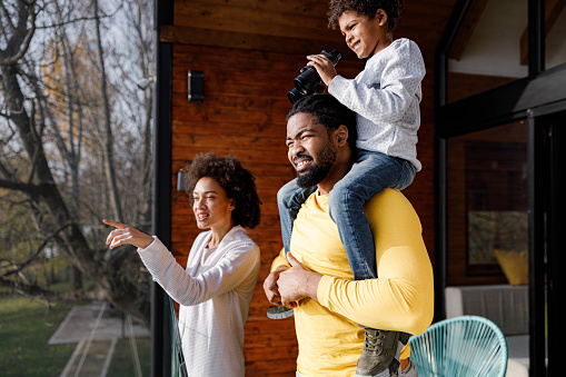 Happy African American family enjoying on a terrace while mother is aiming at distance and son is looking at it through binoculars from his father's shoulders.