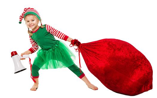 Christmas Kid pulling Santa Bag Full of Gifts. Funny Girl in Elf Costume with Red Huge Sack packed Xmas Presents. Happy Christmas Child with Megaphone. Winter Shopping and Package Delivery isolated White