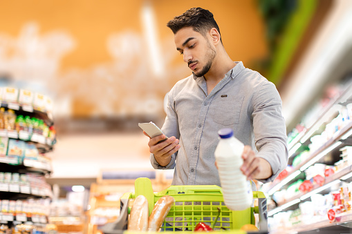 Grocery Shopping Application. Arabic Guy Using Cellphone Holding Milk Bottle Buying Groceries Products Standing With Shop Cart In Supermarket. Consumerism And Technology Concept