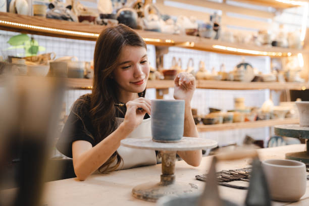 Beautiful young woman holding pottery instrument for scraping, smoothing, shaping and sculpting. Lady siting on bench with pottery wheel and making clay pot stock photo