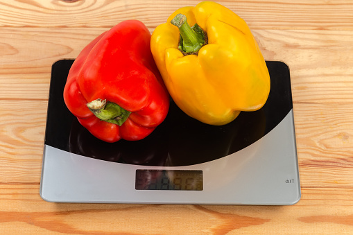 Two whole ripe yellow and red bell peppers on the household digital kitchen scale on the rustic table