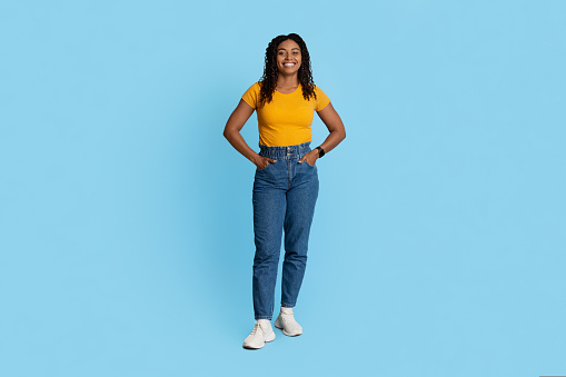 Pretty stylish young african american lady in casual outfit posing alone on blue studio background, happy millennial black woman with curly hair, panorama with copy space, full length