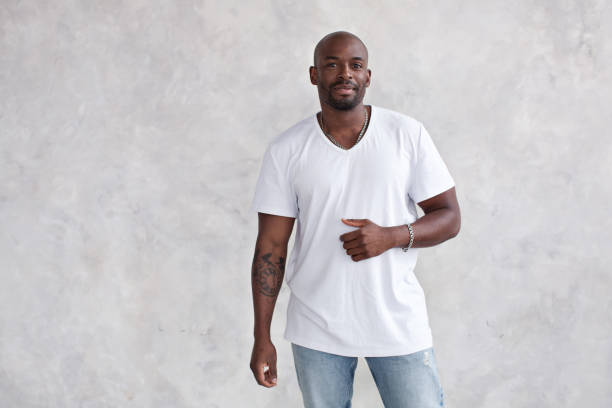 istock Handsome African American young man with tattoo looking at camera, against bright textured wall in studio. Male model wears casual clothes white T-shirt and jeans 1432784814
