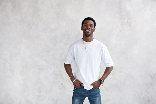 Portrait of smiling African American young man against bright textured wall in studio. Happy male model keeps hands in pockets and wears casual clothes white T-shirt, earrings and chains