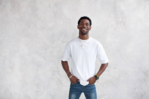 Portrait of smiling African American young man against bright textured wall in studio. Happy male model keeps hands in pockets and wears casual clothes white T-shirt, earrings and chains stock photo