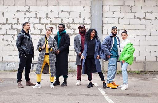 Multiethnic group of young hipsters stands together on street against gray brick wall. Diverse Stylish people in outerwear outdoor, youth culture