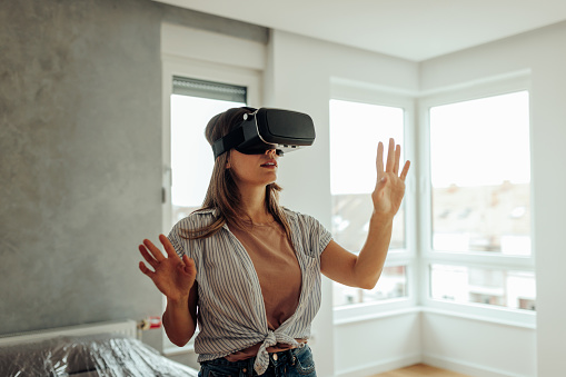 A young woman is using a virtual headset in her new apartment. She is using a virtual layout of her new apartment to better understand on how to position her new furniture.