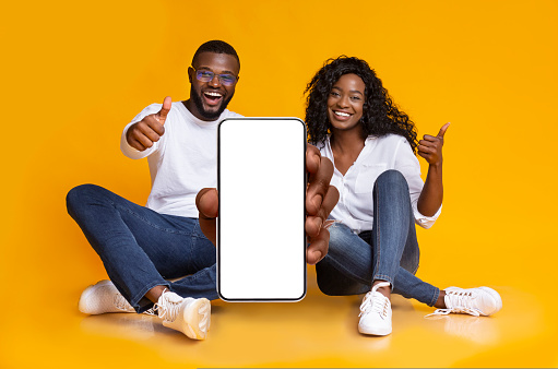 We recommend. Happy black couple demonstrating smartphone with white blank screen and showing thumb up gesture, yellow studio background. Gadget with copy space for mockup