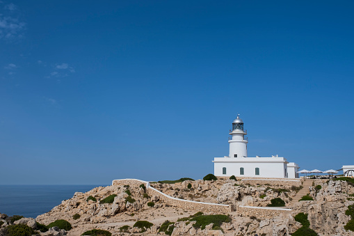 white lighthouse on top of a rocky cliff with white walls surrounding it and the sea in the background, Cavalleria cape lighthouse, Fornells, Menorca Spain, horizontal