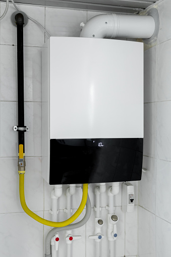 Household budget water heater hanging on the wall in boiler room. Modern gas tanked boiler in bathroom. Common electric storage tank water heater. Energy-efficient home heating system on white tiles