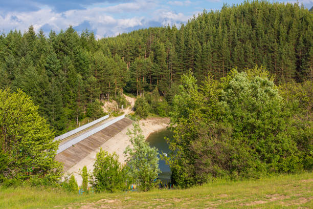 Drone shot of a dam controlling turquoise lake water beside dense green forest. Infrastructure meets nature with winding road and rocky terrain. Scenic landscape with water management and wilderness.