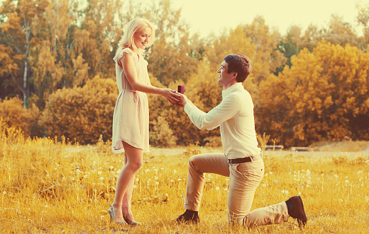 Happy young couple, man kneeling down and proposing a ring to his beloved woman outdoors in the park, wedding concept