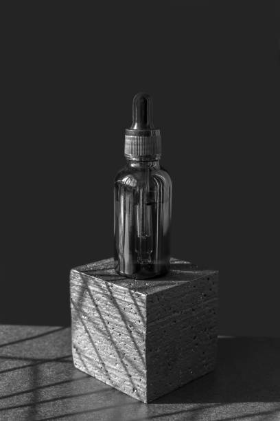 istock Serum in glass bottle with dropper lid on black polystyrene platform. Essential oil for care of women's skin on dark isolated background, in rays of sunlight. Close-up, vertical frame 1432805761