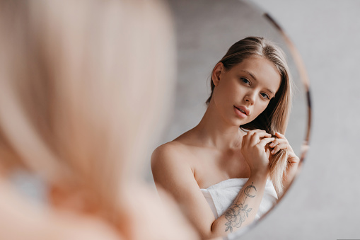 Natural beauty. Pretty young lady touching her soft hair, standing near mirror in bathroom interior and looking at her reflection, over shoulder view