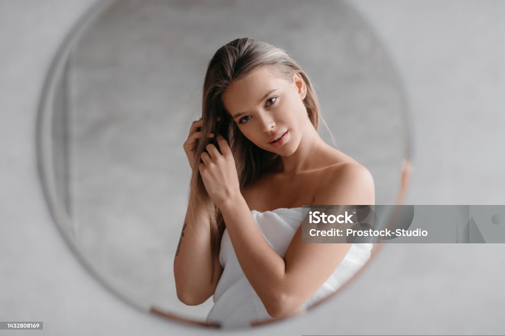 Haircare concept. Young lady in towel touching hair while standing in bathroom and looking at her reflection Haircare concept. Young lady in towel touching hair while standing in bathroom and looking at her reflection. Woman enjoying beauty routine caring for herself Women Stock Photo
