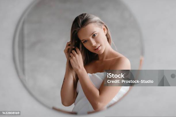 Haircare Concept Young Lady In Towel Touching Hair While Standing In Bathroom And Looking At Her Reflection Stock Photo - Download Image Now