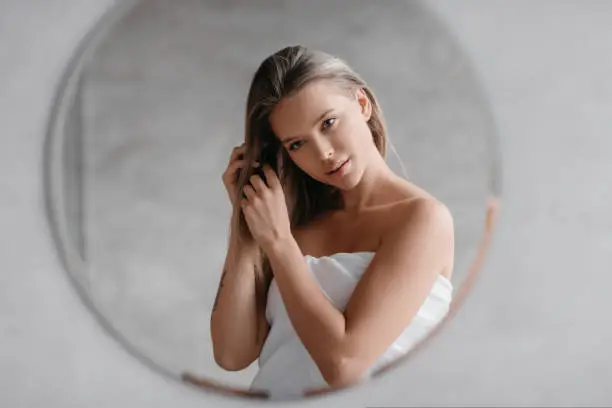 Haircare concept. Young lady in towel touching hair while standing in bathroom and looking at her reflection. Woman enjoying beauty routine caring for herself
