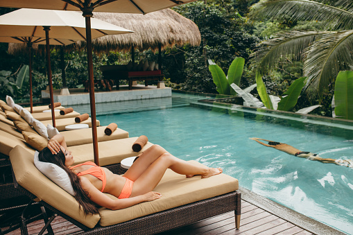 Woman in a bikini relaxing on a lounger next to a resort swimming pool. Serene young woman enjoying a luxury vacation with her man.