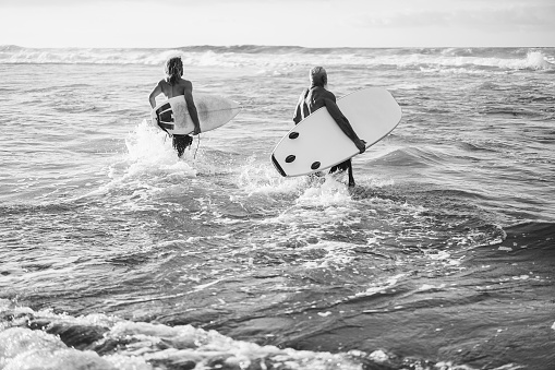 Father and son running on the beach at sunset for surf training - Focus on senior man holding longboard - Black and white editing.