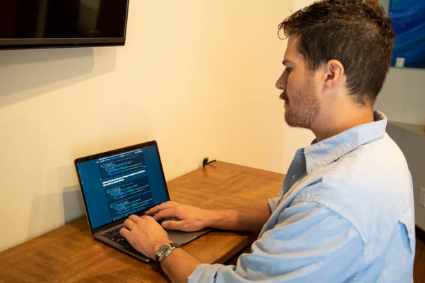 Young man uses laptop while doing web development work He works in a home office and is illuminated by the computer screens cc tv stock pictures, royalty-free photos & images