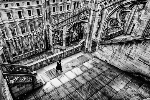 A woman walks towards one of the massive flying buttress and ornately carved stonework on the roof of the Duomo Milano (Milan Cathedral), Italy