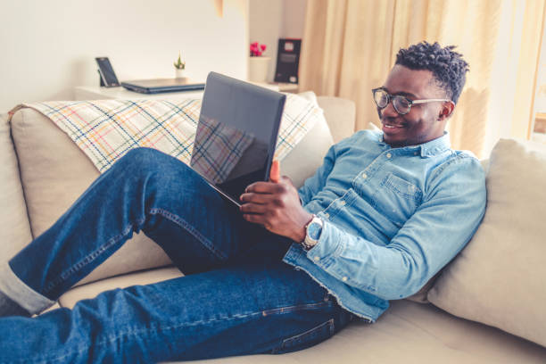 istock Young Afro-american man sitting on couch with laptop, working remotely, using apps. 1432832582