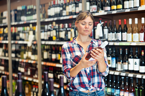 Young woman carefully reads the label on bottle while choosing wine in a supermarket