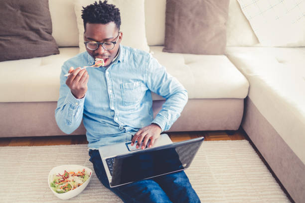istock Young man relaxing at home, sitting on the floor eating a takeaway meal while working on the laptop at home during the day. 1432840574