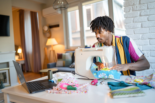 While sitting closely to a sewing machine and his hands on the fabric, a multi-racial man is watching the screen of his laptop open on the table where he is learning a new creative hobby during lockdown