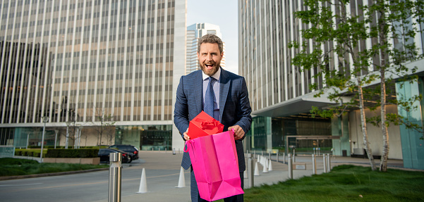 surprised confident entrepreneur in businesslike suit holding shopping bag and giftbox outside the office, boxing day.