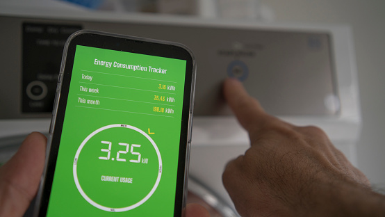 A man is located in a household laundry with laundry appliance in the background. He is holding a smart phone which is displaying current energy consumption