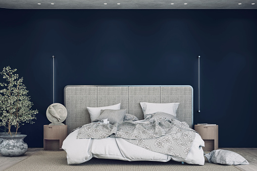 Luxury bedroom with a gray textile headboard bed, white and gray sheets - bedding,  night tables, a sculpture, pillows, a beige carpet, and vertical wall lights on the hardwood floor. A large eucalyptus plant in a pot in front of a rounded navy blue empty plaster wall with copy space and built-in reflector lights on the ceiling.  Front view. 3D rendered image.