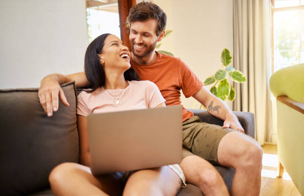 istock Couple talking and browsing on a laptop while relaxing on a sofa at home. Girlfriend and boyfriend scrolling on social media or the internet with technology while having conversation in living room. 1432858959