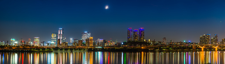 Crescent moon waxing over the neon night skyscrapers and crowded high rise cityscape of Gangnam reflecting in the tranquil waters of the Han River in the heart of Seoul, South Korea’s vibrant capital city.