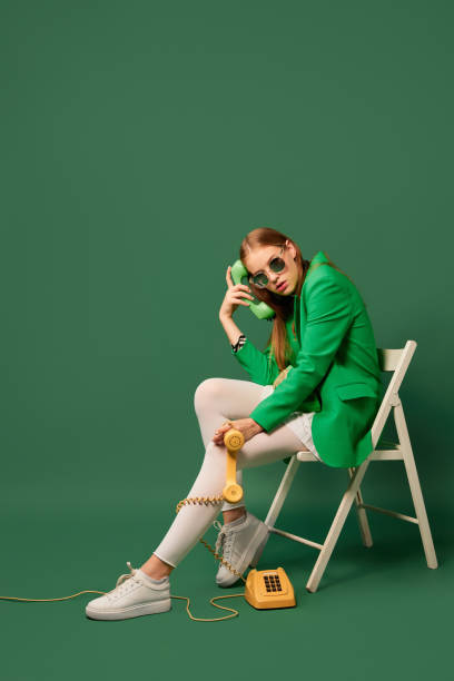 Creative portrait of young without emotions girl in green jacket sitting on chair with retro phone isolated over green background. Vintage, retro style Creative portrait of young without emotions girl in green jacket sitting on chair with retro phone isolated over green background. Vivid style, beauty, queer, freak, fashion concept. Copy space for ad monochrome clothing stock pictures, royalty-free photos & images